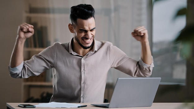 Jonge succesvolle vrolijke Spaanse man mannelijke investeerder zakenman houdt document kijk naar laptop win