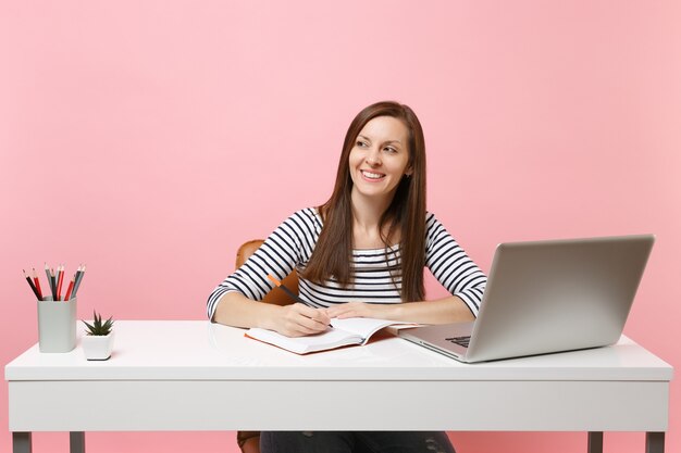 Jonge succesvolle vrouw die opzij kijkt en notities schrijft op een notebook, werkt aan een wit bureau met een moderne pc-laptop geïsoleerd op een pastelroze achtergrond. Prestatie zakelijke carrière concept. Ruimte kopiëren.