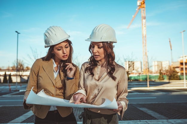 Jonge succesvolle vrouwelijke ingenieurs met witte helmen en blauwdrukken in hun handen analyseren nieuw project op de bouwplaats.