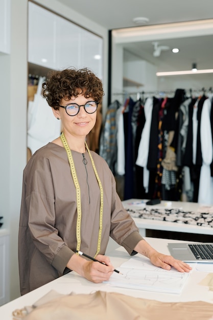 Jonge succesvolle vrouwelijke kleermaker die naar je kijkt tijdens het schetsen op papier door het bureau in haar atelier