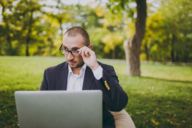 Jonge succesvolle zakenman in wit overhemd, klassiek pak, corrigeert handbril. Man zit op zachte poef, werk op laptop pc-computer in stadspark op groen gazon buitenshuis. Mobiel kantoor, bedrijfsconcept.