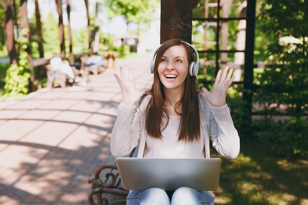 Jonge succesvolle zakenvrouw in lichte vrijetijdskleding. vrouw die werkt op moderne laptop pc-computer, luister muziek in koptelefoon op hoofd in straat buitenshuis. mobiel kantoor. freelance bedrijfsconcept.