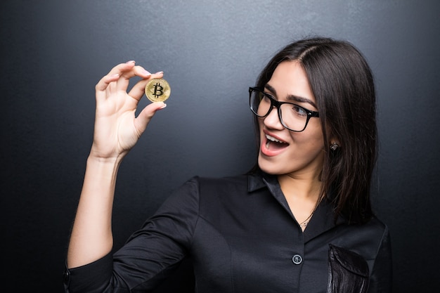 Jonge succesvolle zelfverzekerde vrouw met bril houdt een gouden bitcoin in haar hand geïsoleerd op zwarte muur