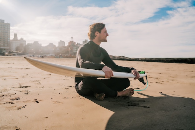 Jonge surfer zittend op zandstrand kijken naar de oceaan met zijn surfplank. Sport en watersport concept.