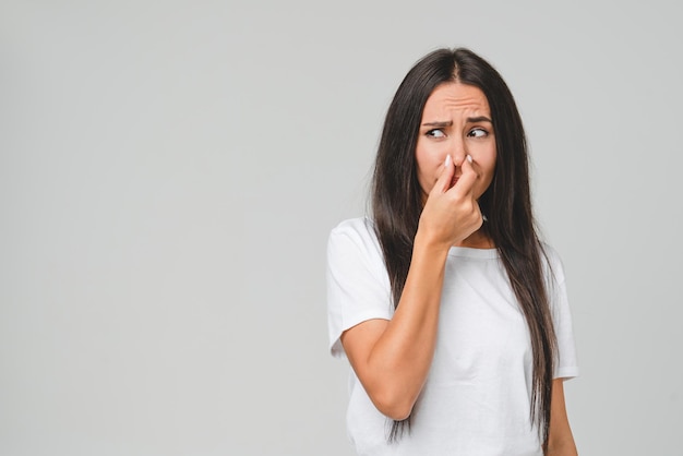 Jonge teleurgestelde verdrietige blanke vrouw in wit T-shirt die stinkend ruikt en haar neus met een hand sluit vanwege de geur die geïsoleerd is op een grijze achtergrond.