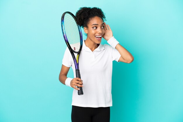 Jonge tennisser vrouw geïsoleerd op blauwe achtergrond luisteren naar iets door hand op het oor te leggen