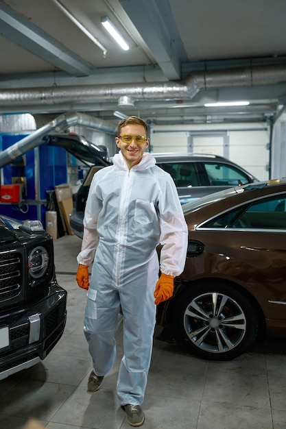 Jonge tevreden verfmeester in veiligheidskleding omringd door gerenoveerde auto. Autogarageservice