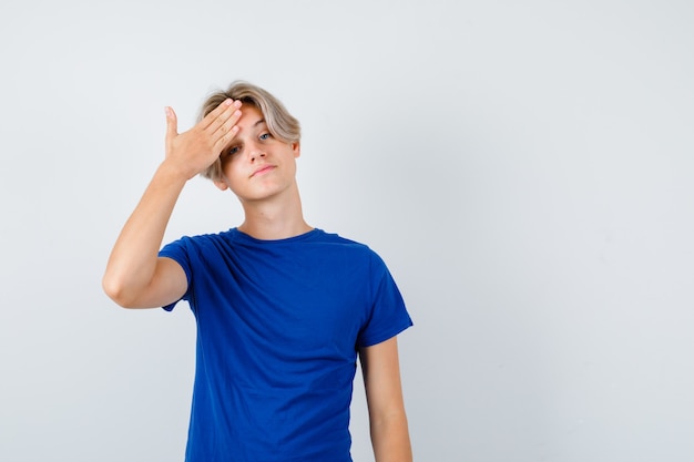 Jonge tienerjongen in blauw t-shirt met hand op voorhoofd en zelfverzekerd, vooraanzicht.