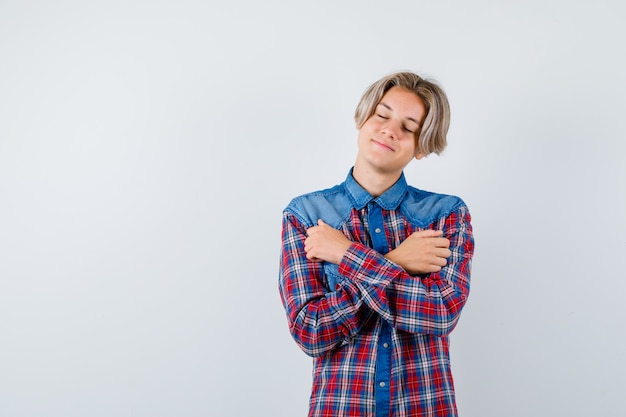 Foto jonge tienerjongen in geruit overhemd knuffelt zichzelf, houdt zijn ogen dicht en ziet er vredig uit, vooraanzicht.