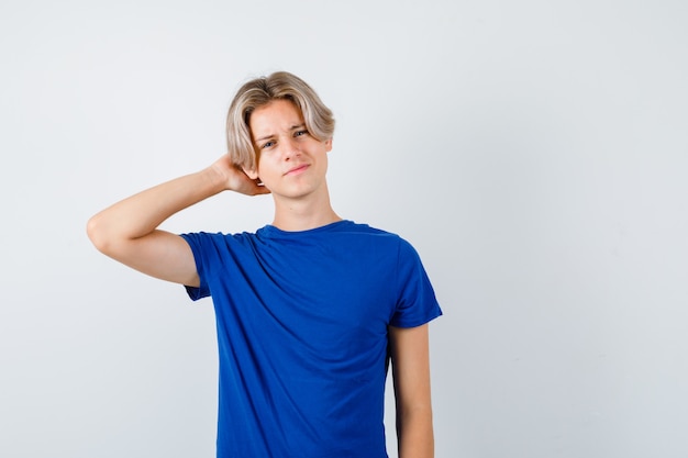 Jonge tienerjongen met hand achter hoofd in blauw t-shirt en peinzend, vooraanzicht.