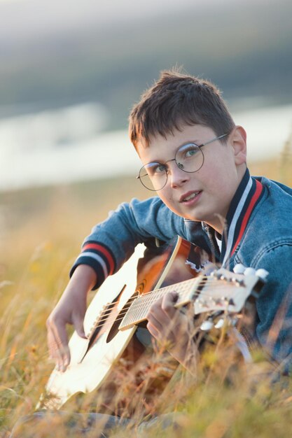 Jonge tienerjongen spelen op akoestische gitaar op zomerveld bij zonsondergang en kijken naar camera