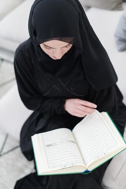 jonge traditionele moslimvrouw die koran leest op de bank voor het iftar-diner tijdens een ramadan-feest thuis