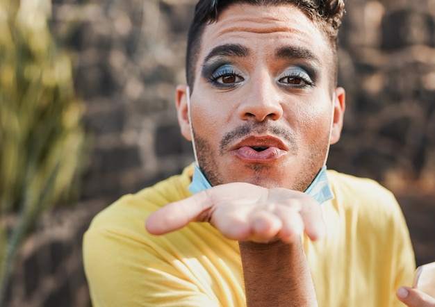 Jonge transgender die kusjes op de camera stuurt terwijl hij een chirurgisch gezichtsmasker onder de kin draagt - Man met make-up buiten