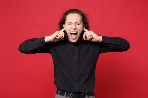 Jonge triest krullend langharige man in zwart shirt staande poseren geïsoleerd op rode muur achtergrond studio portret. Mensen oprechte emoties levensstijl concept. Bespotten kopie ruimte. Probleem crisis depressie