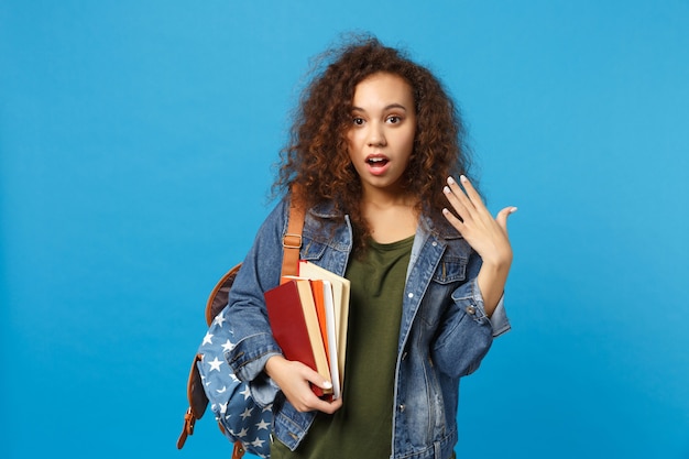 Jonge trieste student in denim kleding en rugzak houdt boeken geïsoleerd op blauwe muur