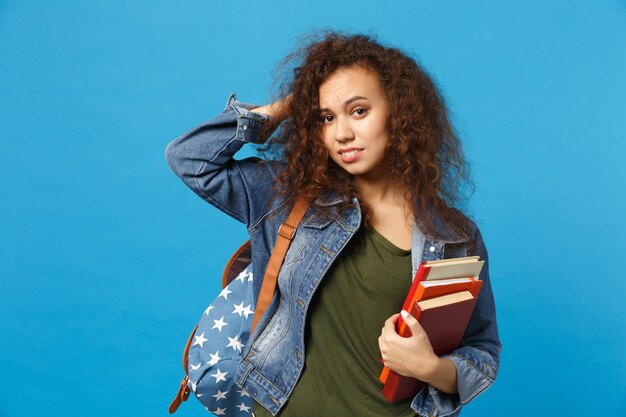 Jonge trieste student in denim kleding en rugzak houdt boeken geïsoleerd op blauwe muur