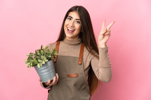 Jonge tuinman meisje met een plant geïsoleerd op roze oppervlak glimlachend en overwinningsteken tonen