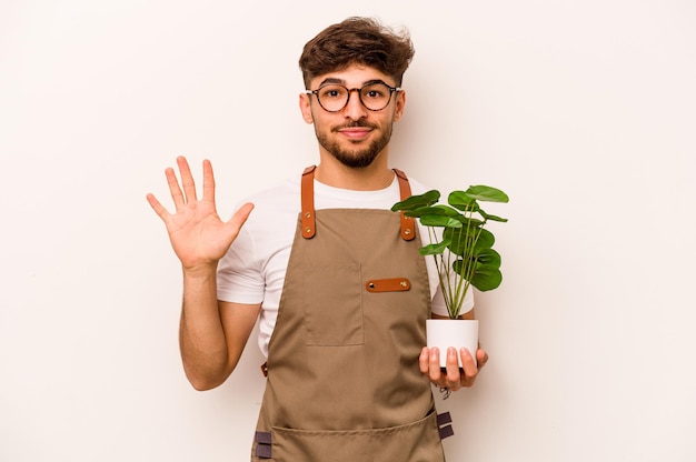 Jonge tuinman Spaanse man met een plant geïsoleerd op een witte achtergrond glimlachend vrolijk met nummer vijf met vingers
