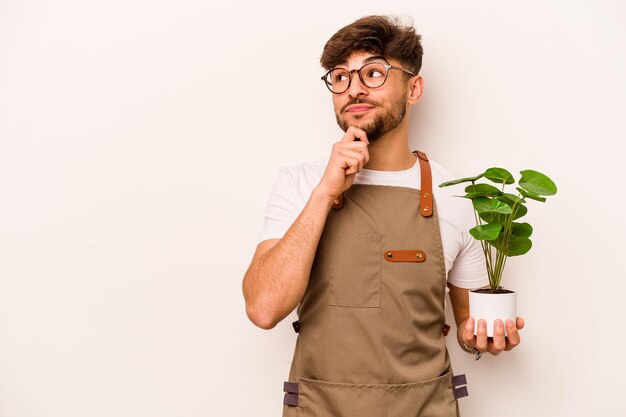 Jonge tuinman Spaanse man met een plant geïsoleerd op een witte achtergrond zijwaarts kijkend met twijfelachtige en sceptische uitdrukking