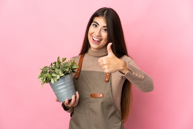 Jonge tuinman vrouw met een plant geïsoleerd op roze muur met duimen omhoog omdat er iets goeds is gebeurd