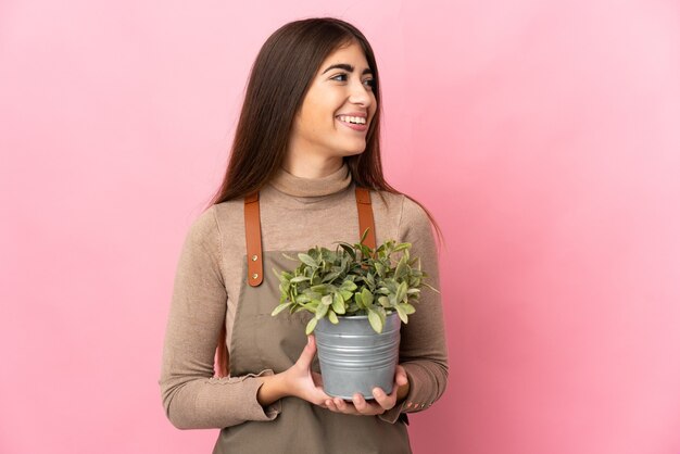 Jonge tuinmanvrouw die een installatie houdt die op roze muur wordt geïsoleerd die kant kijkt