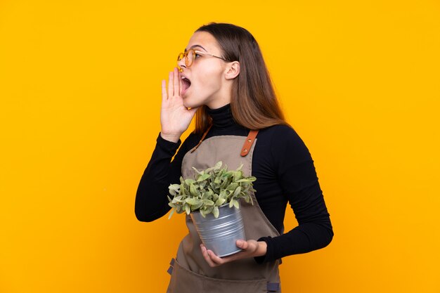 Jonge tuinmanvrouw die een installatie over geïsoleerde gele muur houden die met wijd open mond schreeuwen