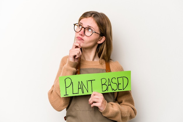 Foto jonge tuinmanvrouw die een op plan gebaseerd plakkaat houdt dat op witte achtergrond wordt geïsoleerd die zijdelings met twijfelachtige en sceptische uitdrukking kijkt.