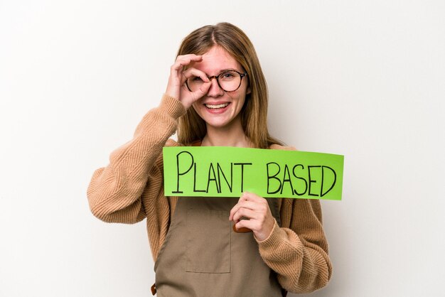 Jonge tuinmanvrouw die een op plan gebaseerd plakkaat houdt dat op witte achtergrond wordt geïsoleerd en opgewonden houdt ok gebaar op oog