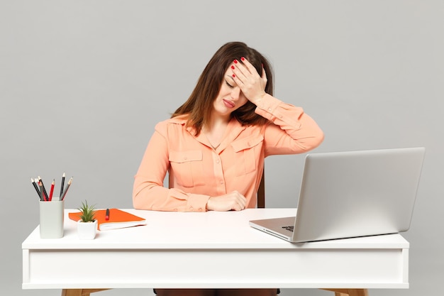 Jonge uitgeputte vrouw in pastel casual kleding hand op het hoofd zitten en werken aan bureau met pc-laptop geïsoleerd op een grijze achtergrond. Prestatie zakelijke carrière levensstijl concept. Bespotten kopie ruimte.