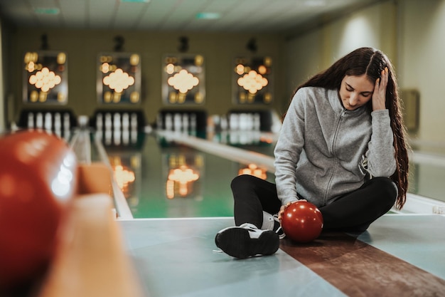 Jonge uitputtingsvrouw zittend op een baan met een bowlingbal en teleurgesteld voelen na het mislukken van het gooien naar de bowlingclub.