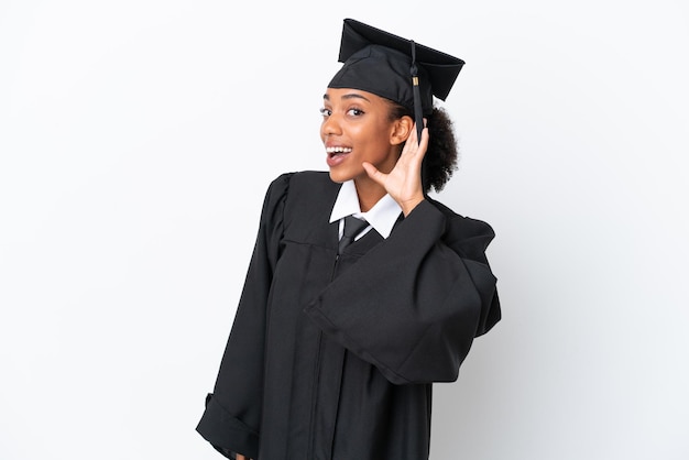 Jonge, universitair afgestudeerde Afro-Amerikaanse vrouw geïsoleerd op een witte achtergrond die naar iets luistert door de hand op het oor te leggen