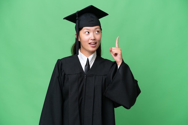 Jonge universitair afgestudeerde Aziatische vrouw over geïsoleerde achtergrond die omhoog wijst en verrast is