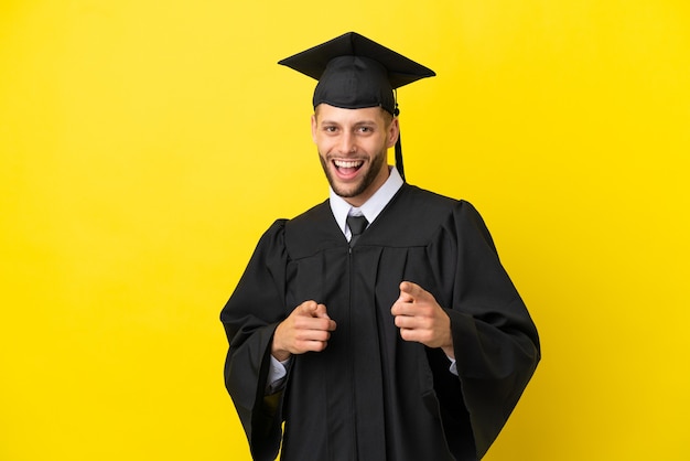 Jonge, universitair afgestudeerde blanke man geïsoleerd op een gele achtergrond die naar voren wijst en glimlacht