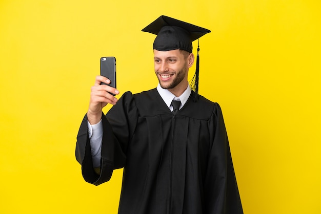 Jonge, universitair afgestudeerde blanke man geïsoleerd op gele achtergrond die een selfie maakt