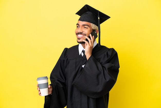 Jonge, universitair afgestudeerde Colombiaanse man geïsoleerd op een gele achtergrond met koffie om mee te nemen en een mobiel