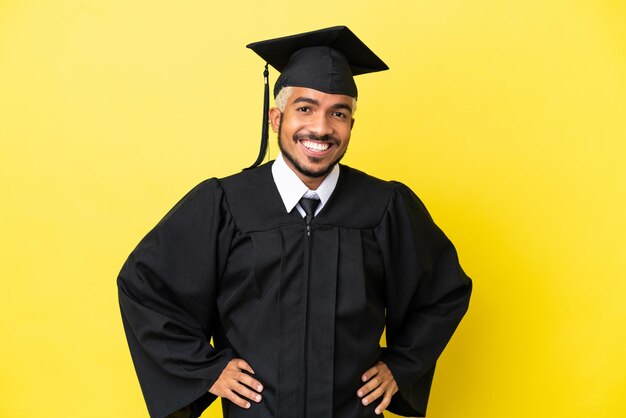 Jonge, universitair afgestudeerde Colombiaanse man geïsoleerd op gele achtergrond poseren met armen op heup en lachend