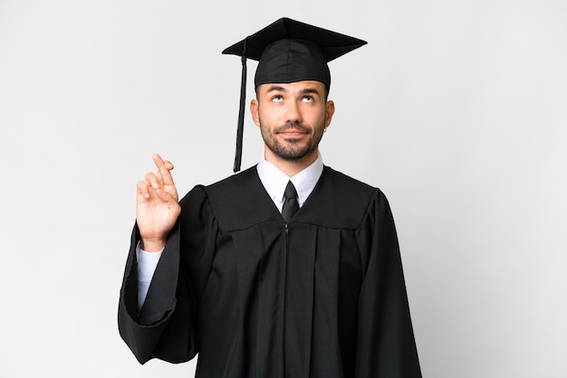 Jonge universitair afgestudeerde man over geïsoleerde witte achtergrond met gekruiste vingers en het beste wensen