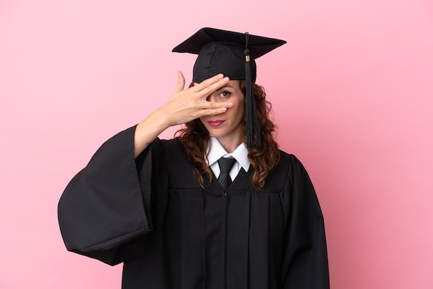 Jonge universitair afgestudeerde vrouw geïsoleerd op roze achtergrond die ogen bedekt door handen en glimlachend