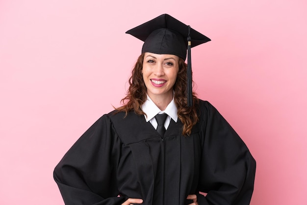 Jonge universitair afgestudeerde vrouw geïsoleerd op roze achtergrond poseren met armen op heup en lachend
