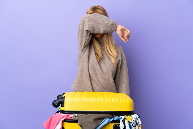 Jonge Uruguayaanse blonde vrouw met een koffer vol kleren over geïsoleerde paarse wandbekleding ogen door handen