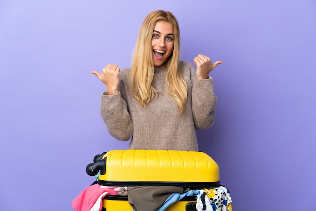 Jonge Uruguayaanse blonde vrouw met een koffer vol kleren over paarse muur met thumbs up gebaar en glimlachen