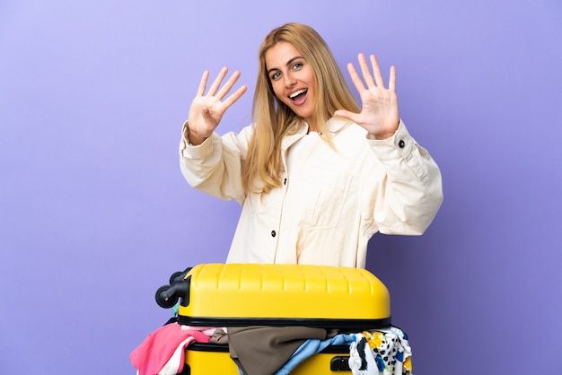 Jonge Uruguayaanse blonde vrouw met een koffer vol kleren over paarse muur tellen negen met vingers