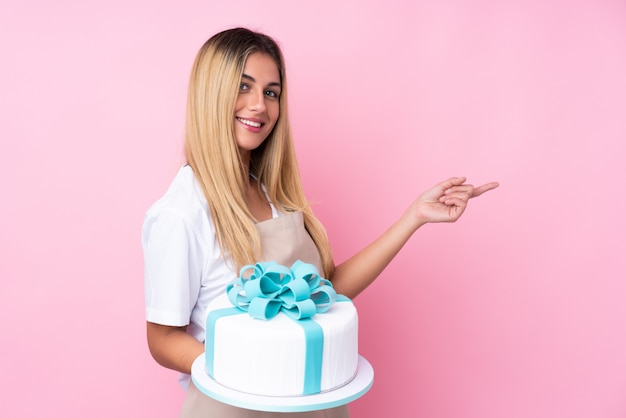 Jonge Uruguayaanse gebakjevrouw met een grote cake over geïsoleerde muur roze muur die aan de kant richt om een product te presenteren