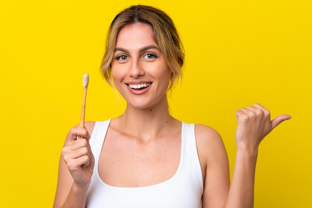 Jonge Uruguayaanse vrouw die tanden poetst geïsoleerd op een gele achtergrond, wijzend naar de zijkant om een product te presenteren