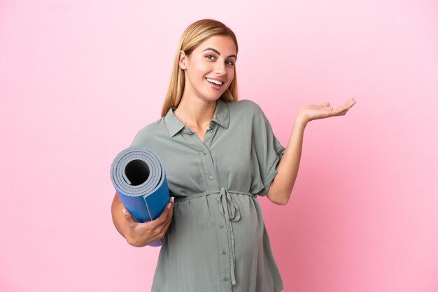 Jonge Uruguayaanse vrouw geïsoleerd op blauwe achtergrond zwanger en houdt een mat vast terwijl ze iets presenteert