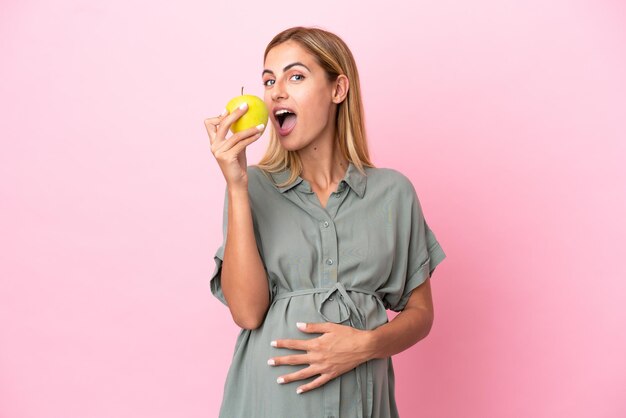 Jonge Uruguayaanse vrouw geïsoleerd op blauwe achtergrond zwanger en met een appel