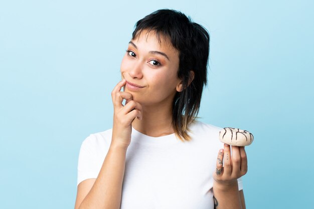 Jonge Uruguayaanse vrouw over geïsoleerde blauwe achtergrond die een doughnut houdt