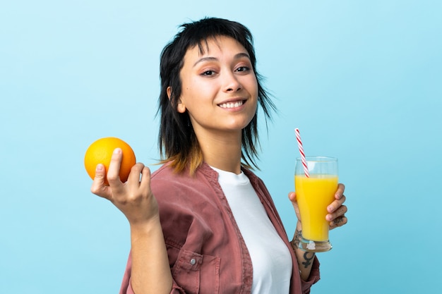 Jonge Uruguayaanse vrouw over geïsoleerde blauwe achtergrond die een sinaasappel en een sinaasappelsap houdt