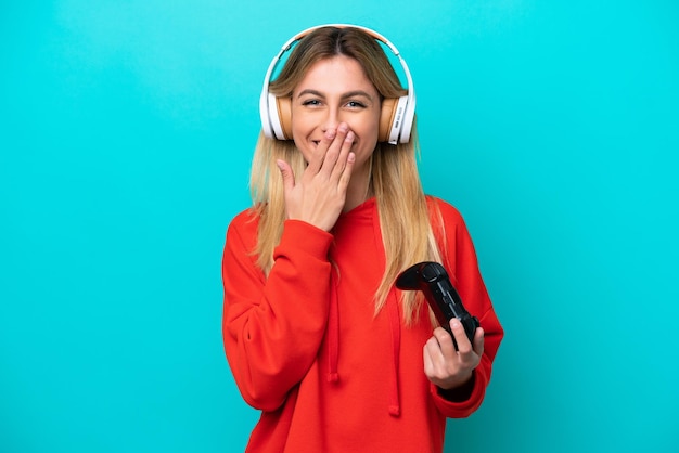 Jonge Uruguayaanse vrouw speelt met een videogamecontroller geïsoleerd op blauw, blij en glimlachend die de mond bedekt met de hand