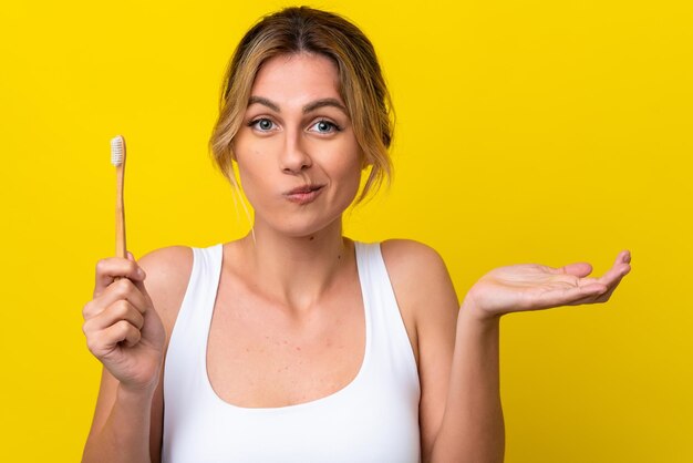 Jonge Uruguayaanse vrouw tandenpoetsen geïsoleerd op gele achtergrond twijfels gebaar maken terwijl ze de schouders optilt